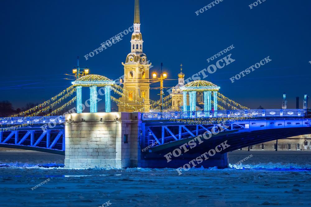 На фотографии изображен мост в Санкт-Петербурге, украшенный праздничной иллюминацией в зимний период. Световые гирлянды подчёркивают его арочные конструкции и создают яркий контраст с темным небом. Вдали видна Петропавловская крепость с её узнаваемой колокольней. Это место, украшенное к Новому году, выглядит особенно эффектно и привлекает множество жителей и гостей города.